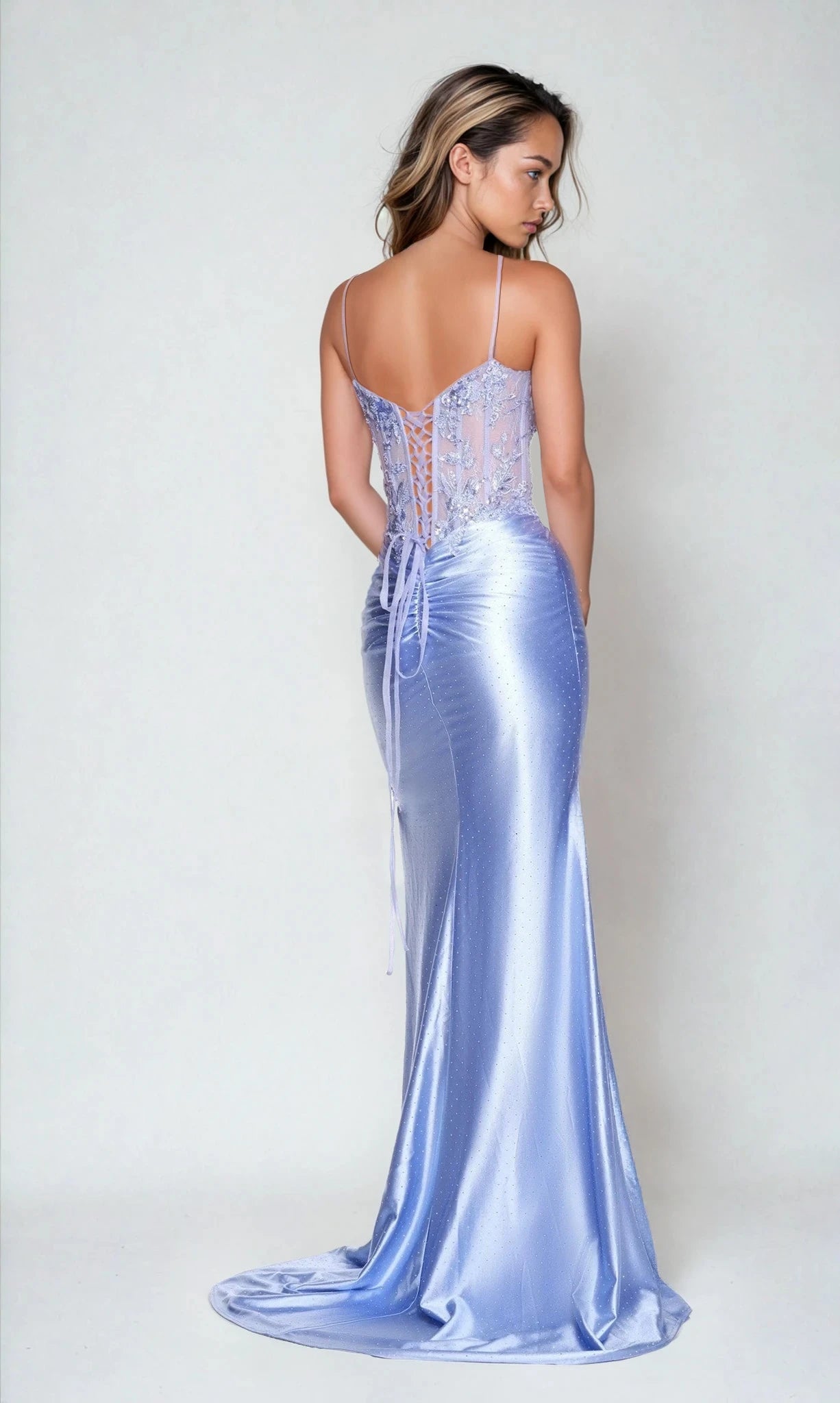 A woman in the Dave and Johnny 12750 Long Prom Dress—light purple satin with a corset bodice, lace-up back, thin straps, and a sleek sheath skirt—stands against a plain light background, facing away from the camera.